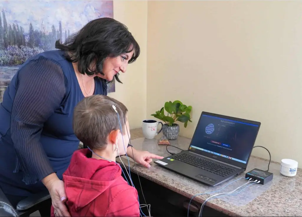 Adult assisting child wearing neurofeedback headset at desk with laptop
