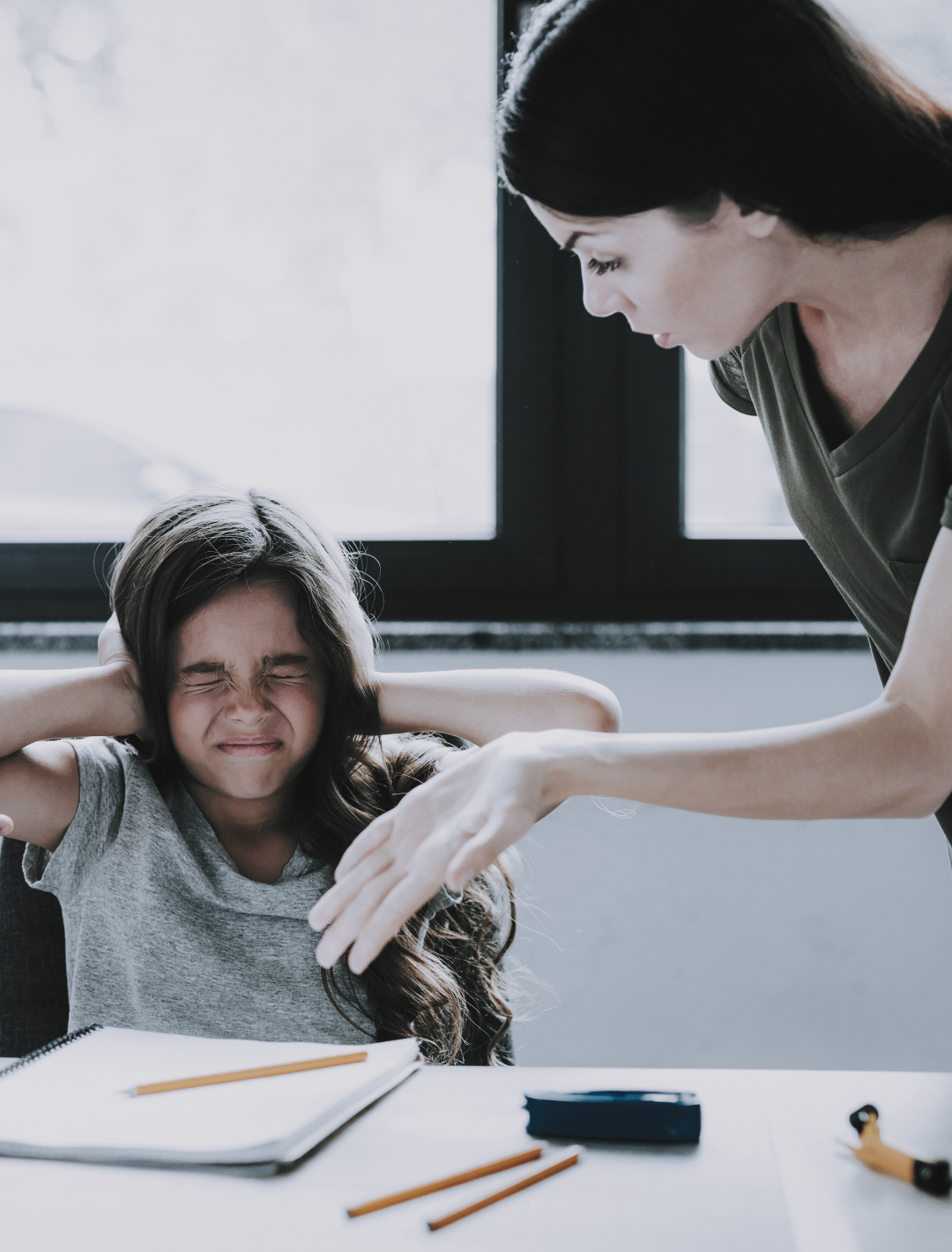 Child with frustrated expression at desk, adult comforting from behind