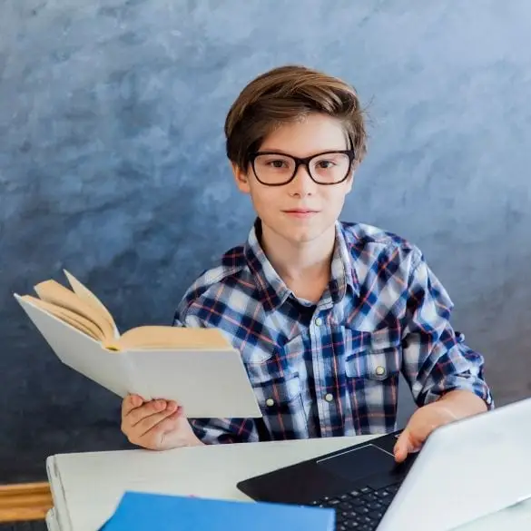 Boy wearing glasses reading a book at desk with laptop