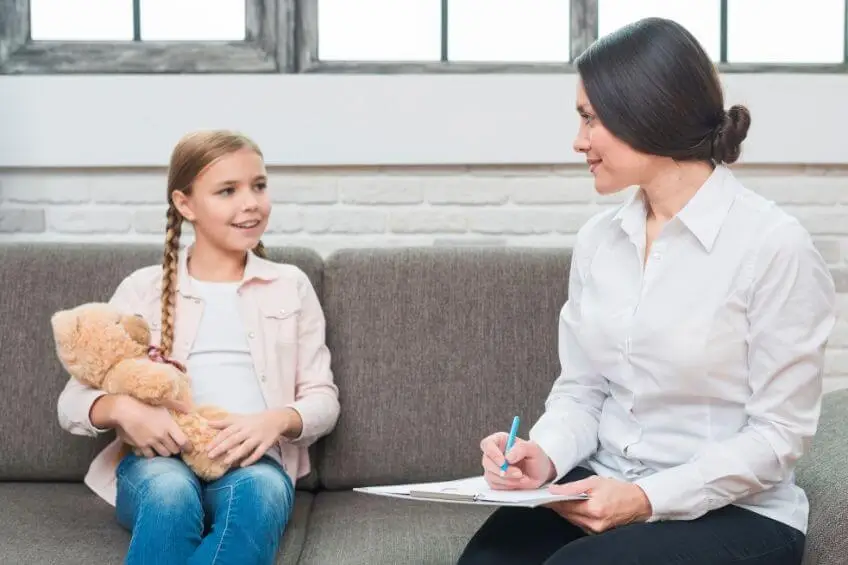 Child holding teddy bear speaking with therapist taking notes on couch