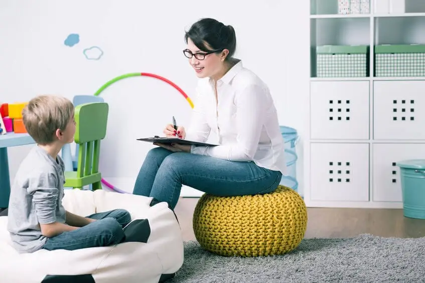 Therapist and child during session in clinical setting with colorful activity materials and storage