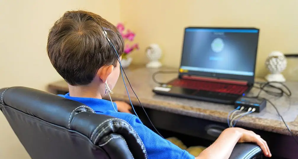 Child from behind at desk with neurofeedback sensors, viewing laptop